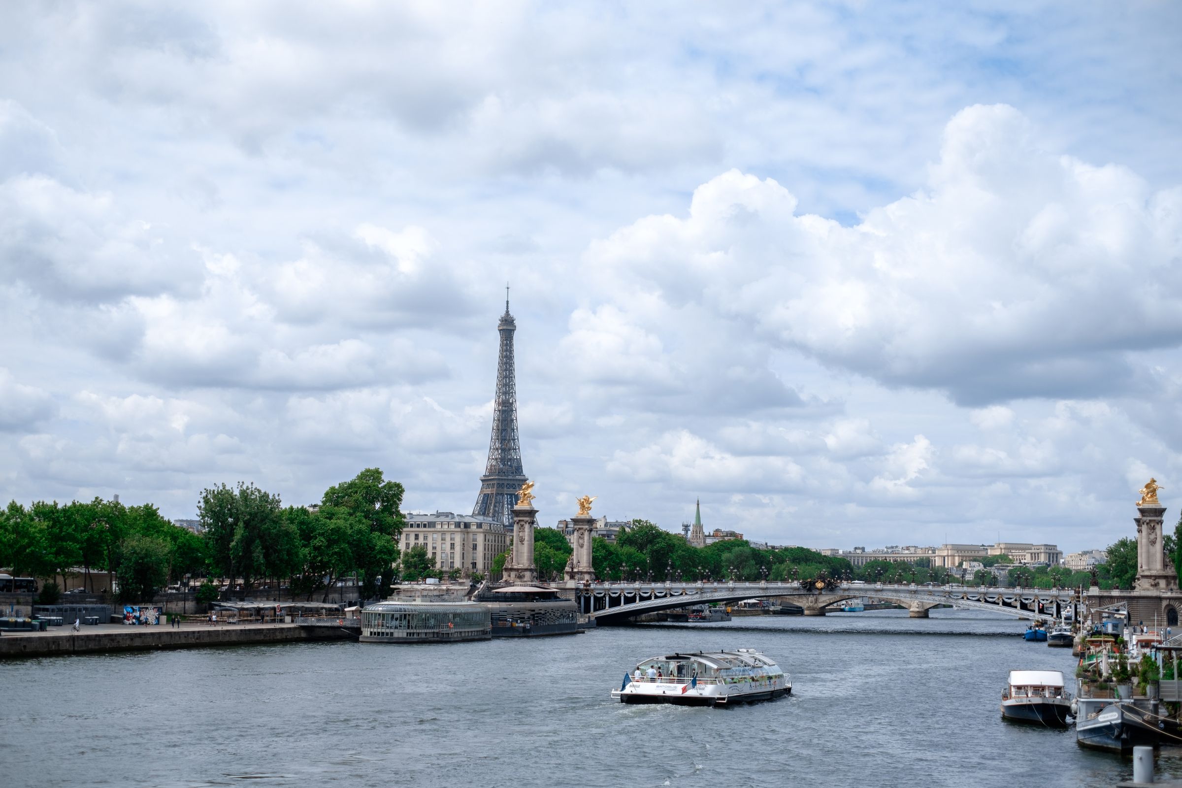 Scenic view of the Eiffel Tower and Seine River in Paris with a bridge and boats under a cloudy sky.